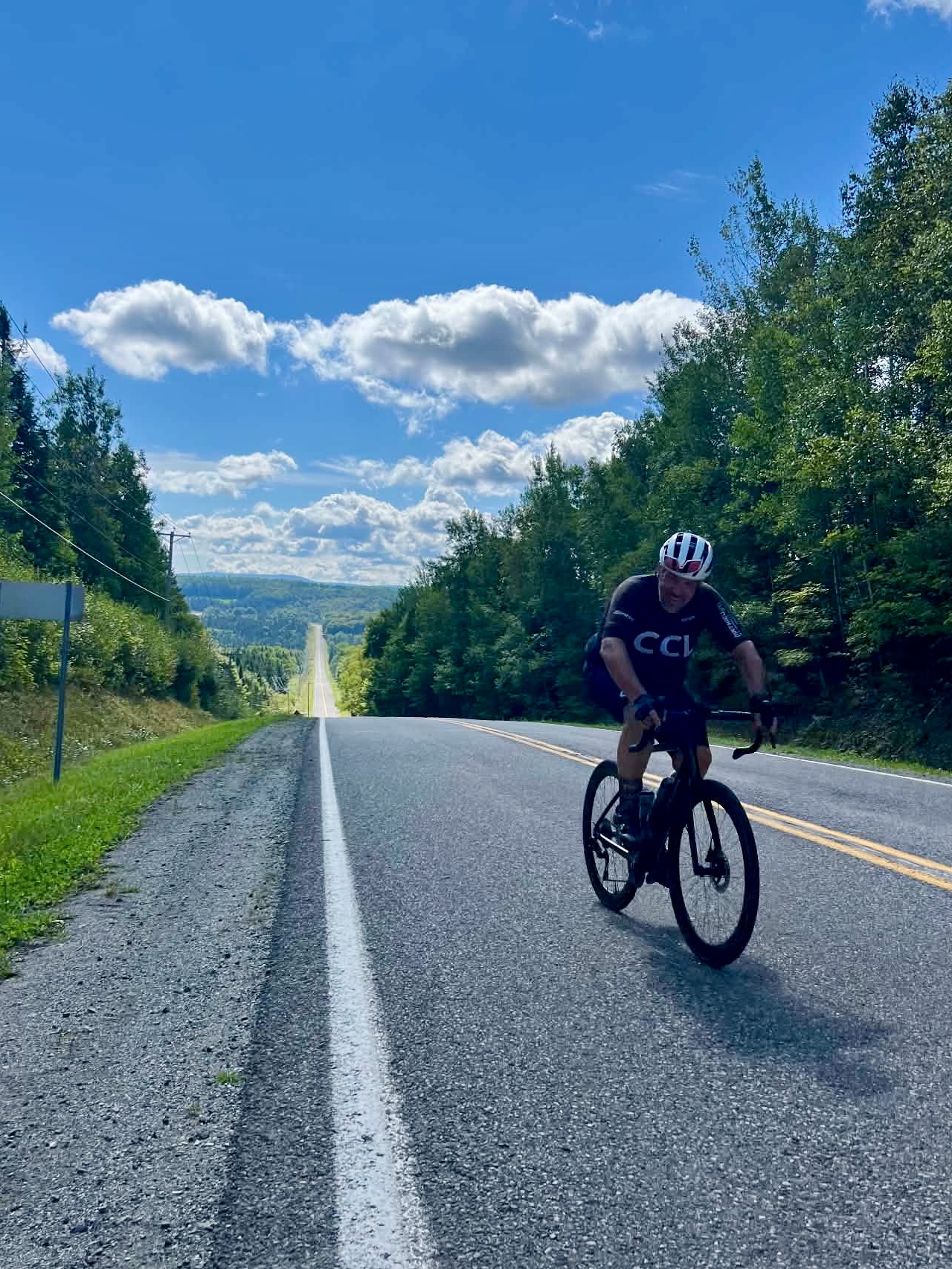 Un cycliste sur une route vallonnée bordée d’arbres, sous un ciel bleu parsemé de nuages